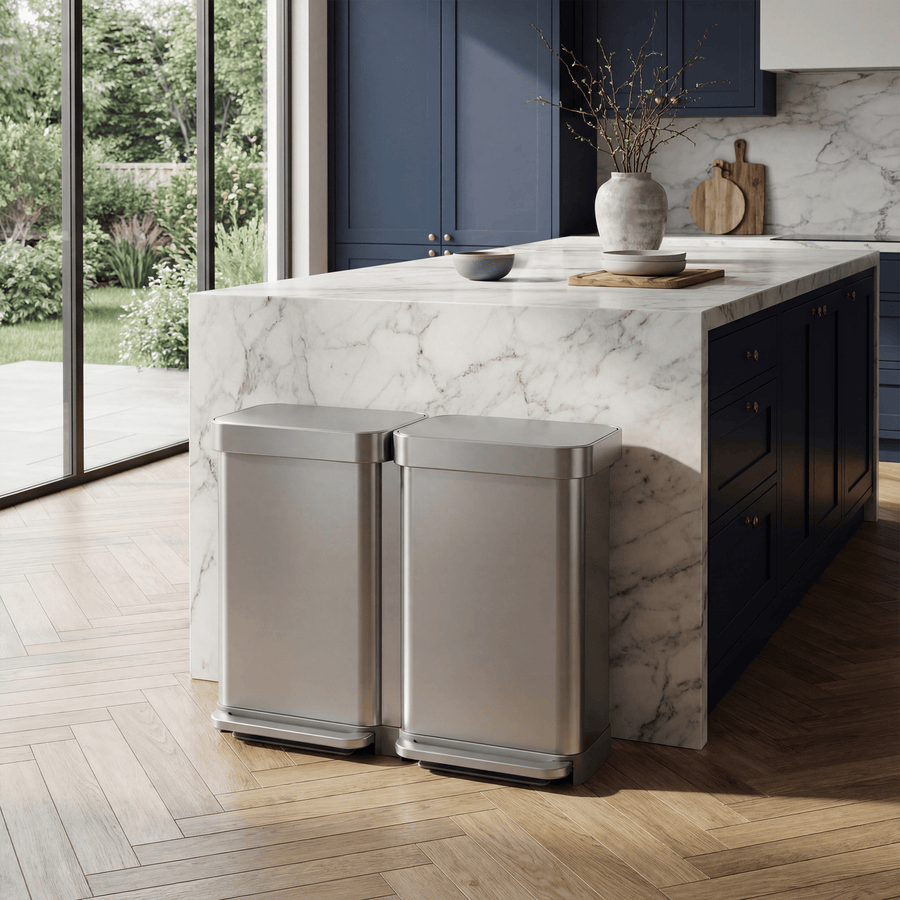 Simplehuman's 90L Step Recycler in Brushed. This image highlights the can in the middle of a lived in modern house. The background features dark blue cabinets, a mix of wooden and marble appliances with gold hardware and few plants. 